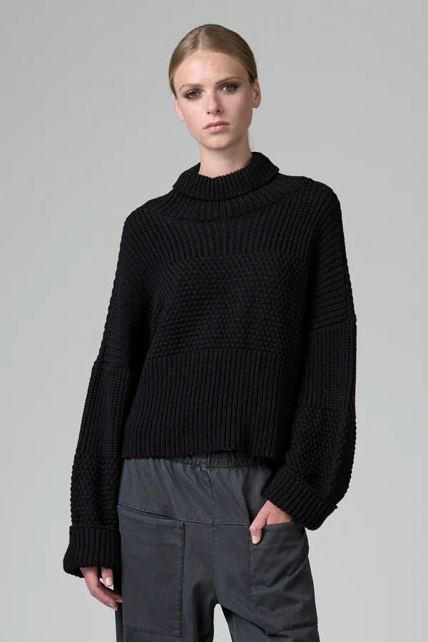 Front view of black wool chain stitch knit ribbed turtleneck on model wearing grey pants, against a grey background.