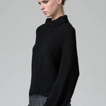 Side view of black wool chain stitch knit ribbed turtleneck on model wearing grey pants, against a grey background.