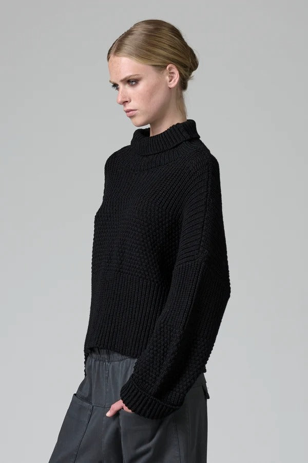 Side view of black wool chain stitch knit ribbed turtleneck on model wearing grey pants, against a grey background.