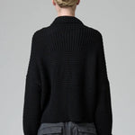 Back view of black wool chain stitch knit ribbed turtleneck on model wearing grey pants, against a grey background.