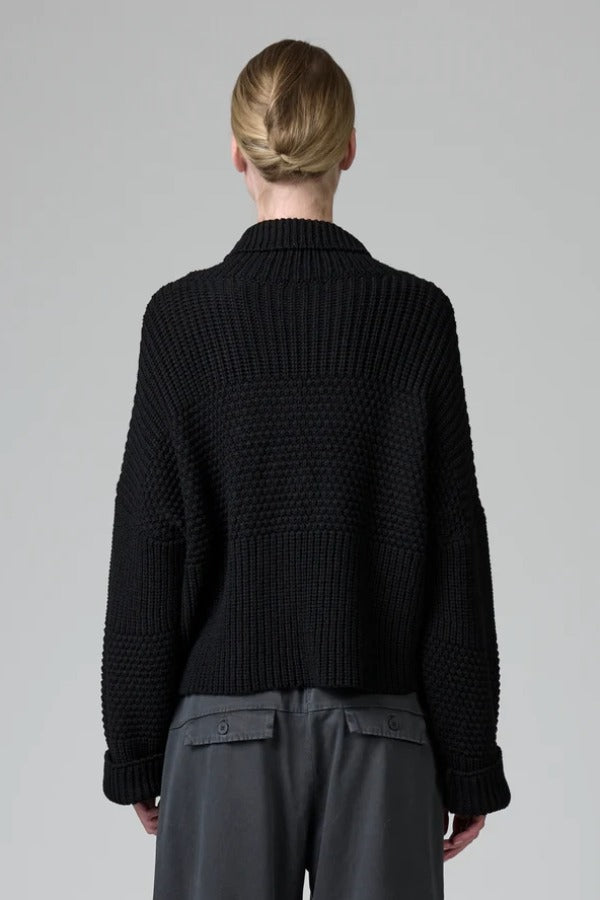 Back view of black wool chain stitch knit ribbed turtleneck on model wearing grey pants, against a grey background.