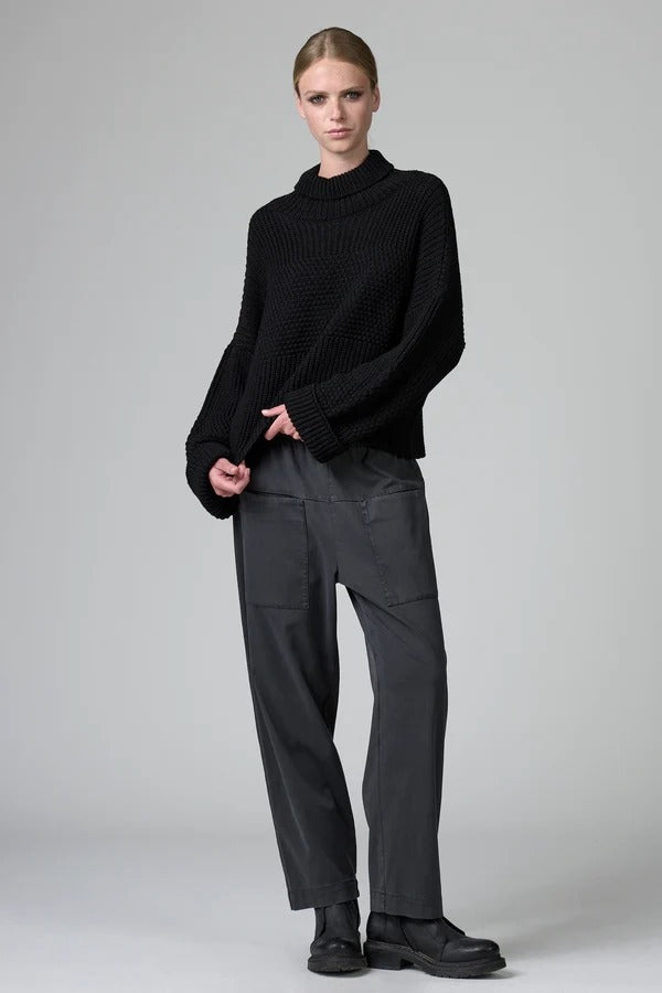 Front view of black wool chain stitch knit ribbed turtleneck on model wearing grey pants, against a grey background.