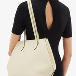 Side view of white valextra's small vivi shopping bag on model wearing black shirt and pants against a white background.