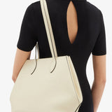 Side view of white valextra's small vivi shopping bag on model wearing black shirt and pants against a white background.