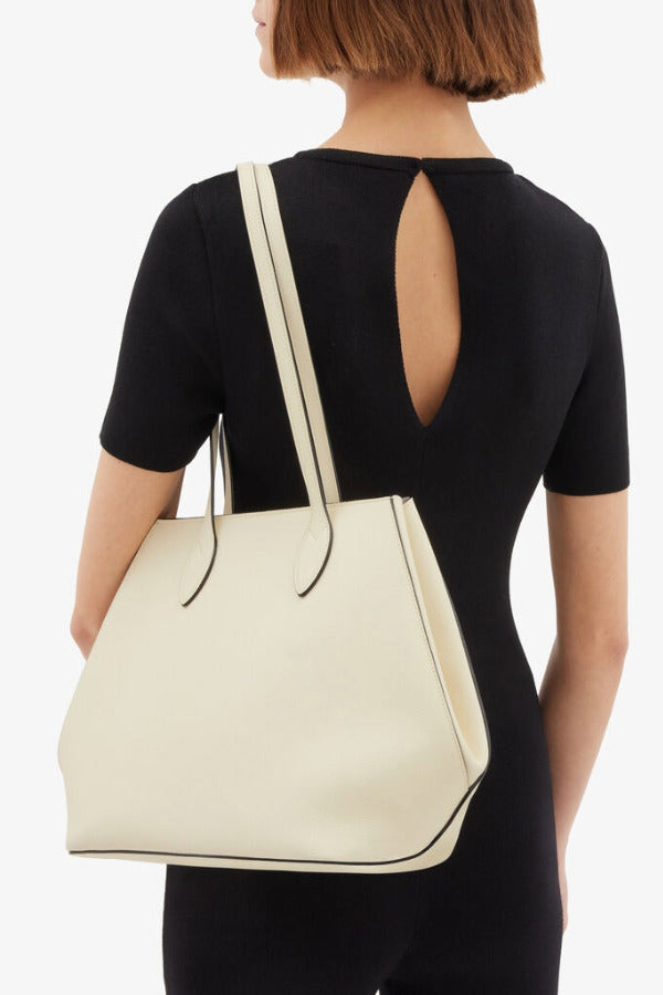 Side view of white valextra's small vivi shopping bag on model wearing black shirt and pants against a white background.