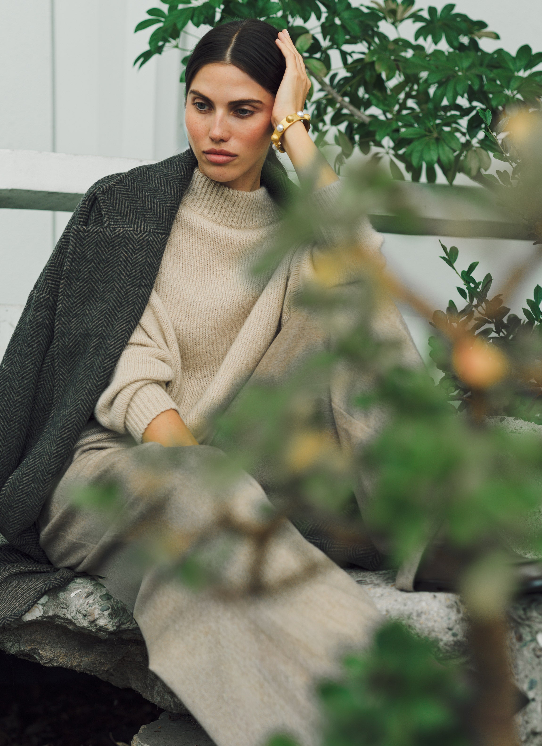 Woman sitting outdoors wearing a beige sweater and gray coat, surrounded by greenery.