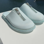 A pair of light blue Guidi zip sandals displayed on a white surface with a marina in the background. The sandals feature a central silver zipper and a textured sole, with yachts and boats visible in the blurred water behind them.