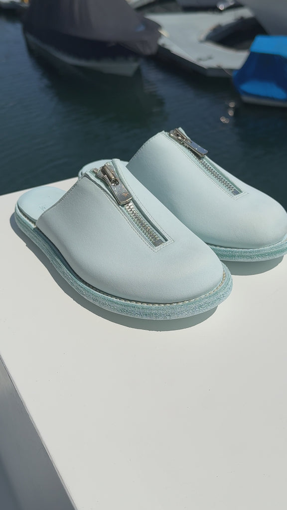 A pair of light blue Guidi zip sandals displayed on a white surface with a marina in the background. The sandals feature a central silver zipper and a textured sole, with yachts and boats visible in the blurred water behind them.