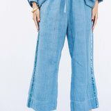 Front closeup view of denim pants with velvet ribbon on model wearing denim tunic and black sandals, against a white background.