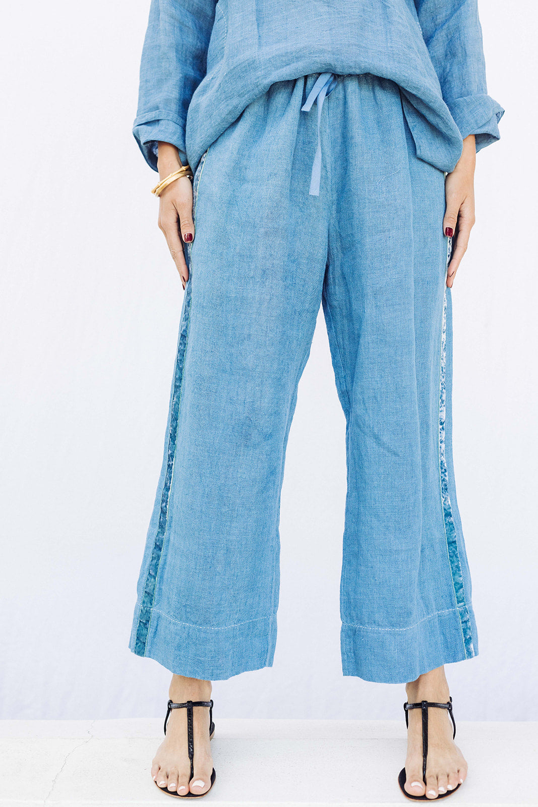Front closeup view of denim pants with velvet ribbon on model wearing denim tunic and black sandals, against a white background.