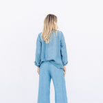 Back view of denim pants with velvet ribbon on model wearing denim tunic and black sandals, against a white background.