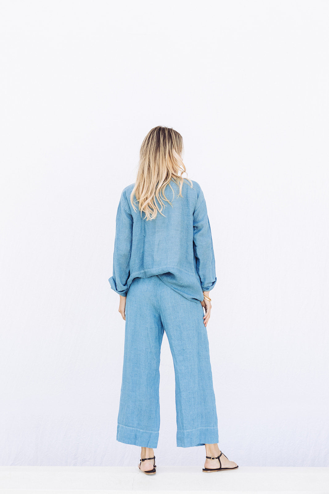 Back view of denim pants with velvet ribbon on model wearing denim tunic and black sandals, against a white background.